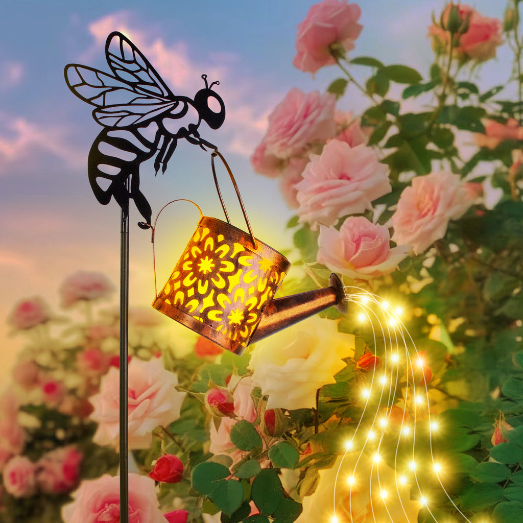 Decorative garden stake with bee and watering can design, surrounded by pink roses and string lights.